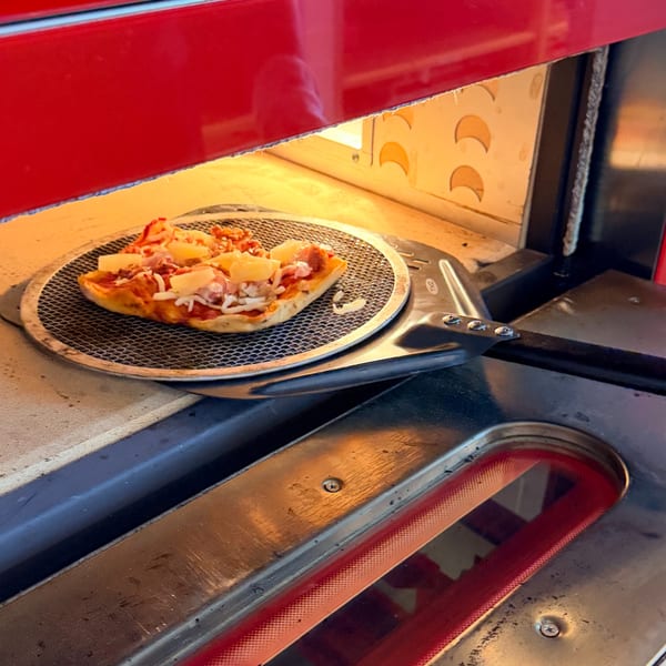 A Hawaiian pizza with pineapple and ham cooking on a metal screen in a red pizza oven.