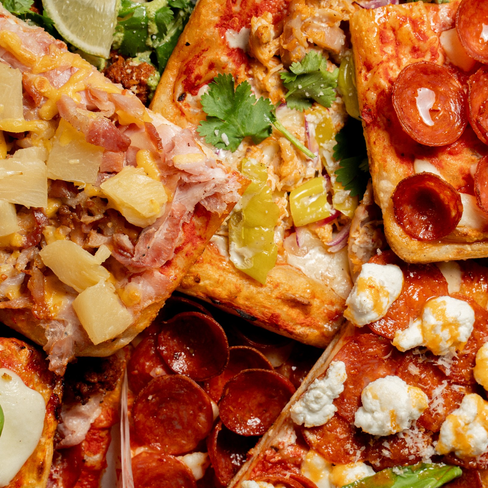 Close-up assortment of pizzas topped with pepperoni, ham, pineapple, tomatoes, peppers, cilantro, and crumbly cheese.