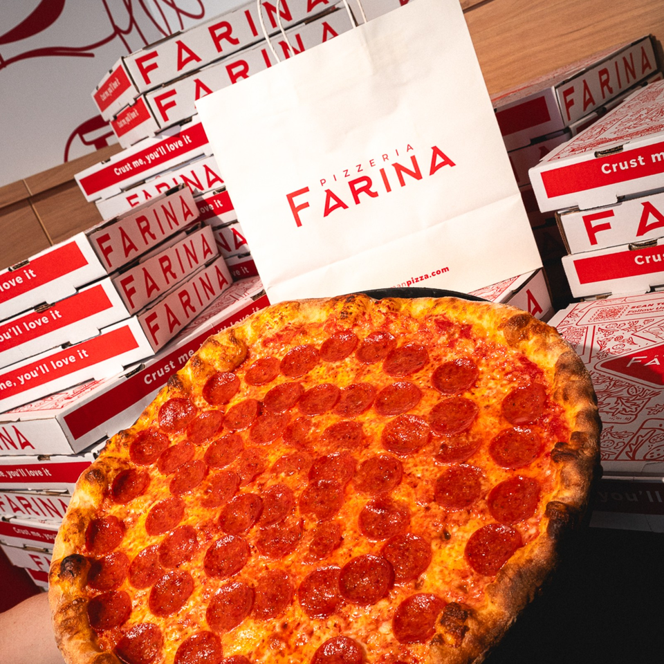 Pepperoni pizza held in front of stacked Farina Pizzeria boxes and branded packaging.